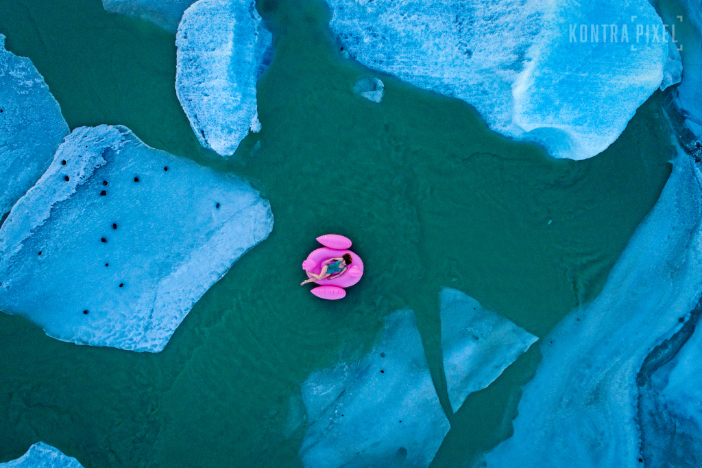 Drohnenaufnahme in Island: Eine Frau liegt auf einem pinkfarbenen Schwimmtier-Pelikan im Badeanzug zwischen Eisschollen im türkisfarbenen Wasser